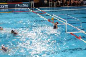 Laura Aarts stopt een bal tijdens een waterpolowedstrijd, gefocust in het doel met gespreide armen in het zwembad.